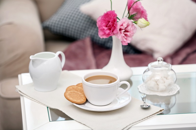 White coffee table with glass top with a cup of coffee, cookies, and cream and sugar, and a vase of flowers