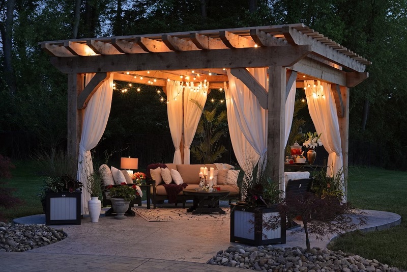 Outdoor backyard oasis in the evening under pergola with lighting, curtains, plants and sofa