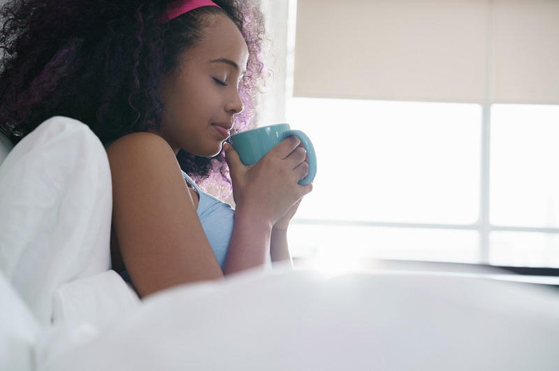 Young woman enjoying a hot cup of coffee early in the morning inside her comfortable bedding