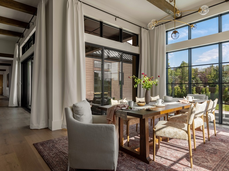 Dining room with a long wooden table, glass chandelier, and floor-to-ceiling windows looking onto trees