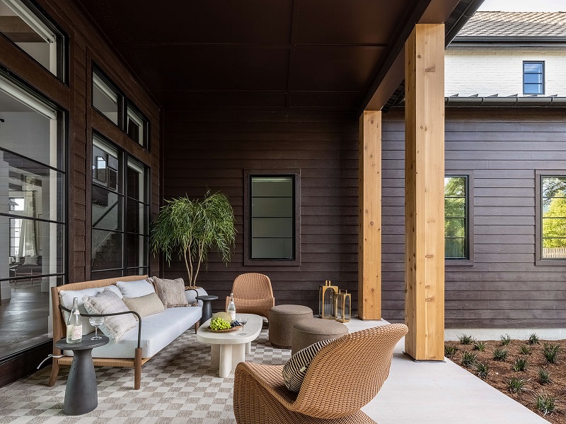 Covered outdoor seating area with a white sofa, woven chairs, wood posts, and glass doors, beside newly planted garden beds